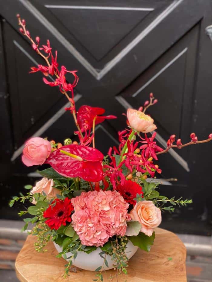 The Watering Can | A Mid-size European Arrangement in a low, round, white ceramic container made up of a variety of flowers in blush and red tones.