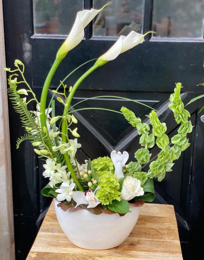 The Watering Can | A mid-size Euopean Arrangement of whites and greens encircling a small angel statue. The arrangemnt rests in a round white ceramic container.