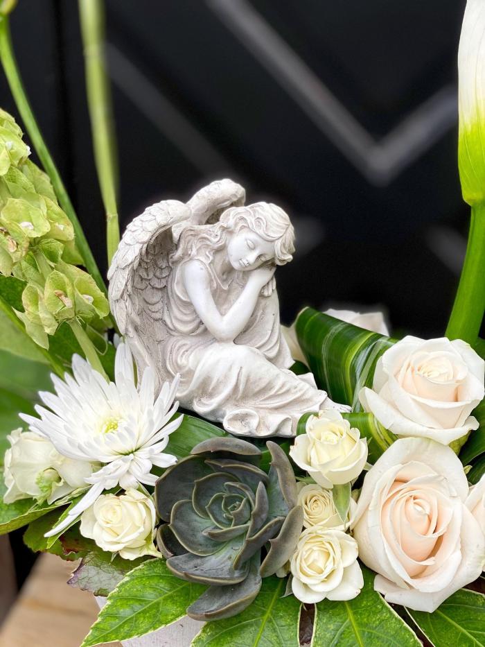 The Watering Can | A close up of a statue of an angel sitting in a bed of white blooms and greens of the flower arrangement.