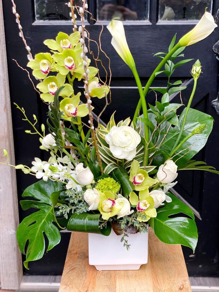 The Watering Can | Stunning large European arrangement featuring a stem of green cymbidium orchids and large white calla lilies emerging from a bed of smaller white flowers and tropical greens.