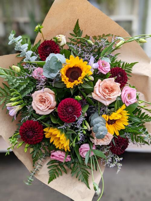 The Watering Can | A hand-tied bouquet freaturing burgundy dahlia, sunflowers, and light pink roses wrapped in brown paper.