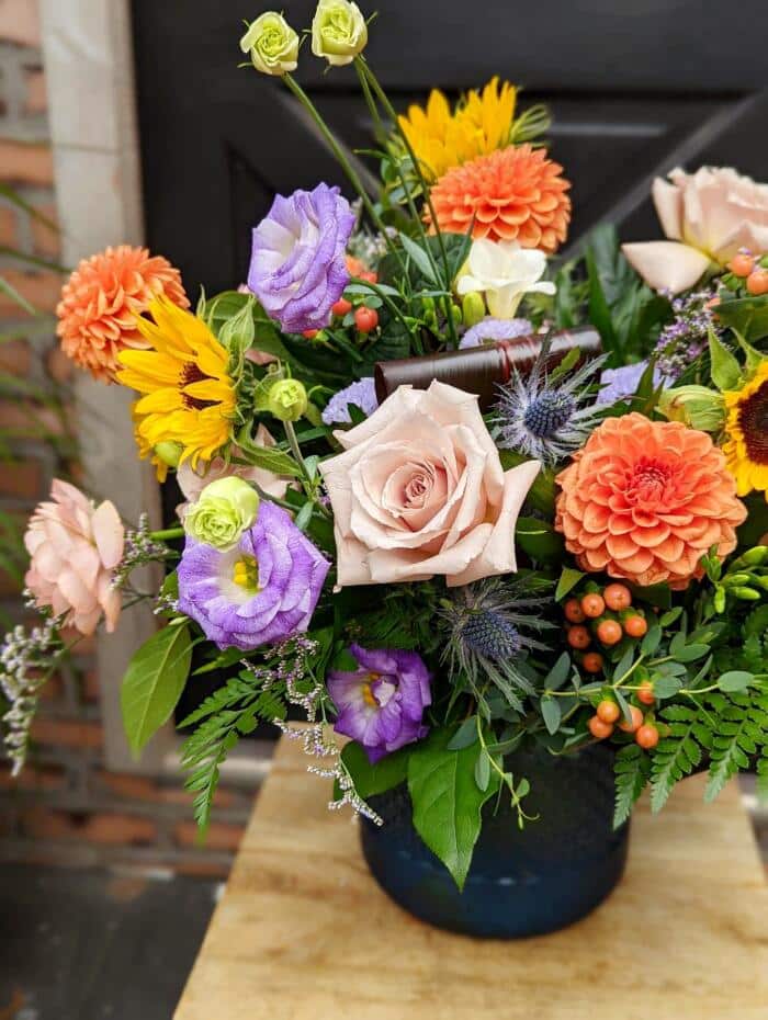 The Watering Can | Orange dahlias, blush roses, purple lisianthus, yellow sunflowers, and peach hypericum beautifully arranged with a variety of greens in a dark blue vase.