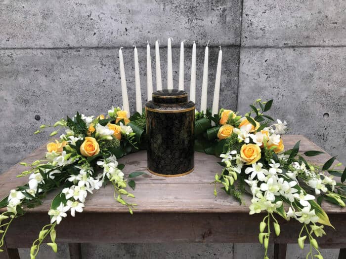 The Watering Can | A semi circular yellow and white floral arrnagement with a row of tall white tapers placed behind the urn.