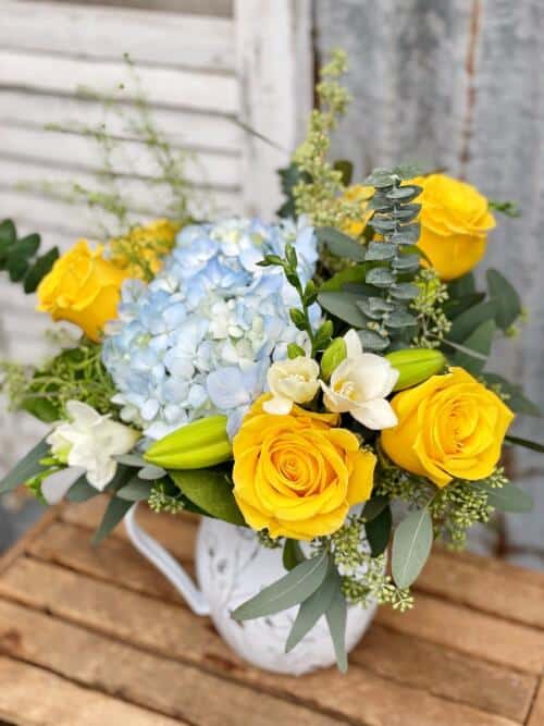 A beautiful bouquet with yellow roses, blue hydrangea and white freesia displayed in a water pitcher
