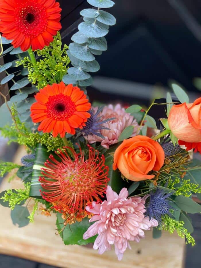 The Watering Can | Red pinchusion, orange roses, orange gerbera daisies and pink mums in a European style floral arrangement.