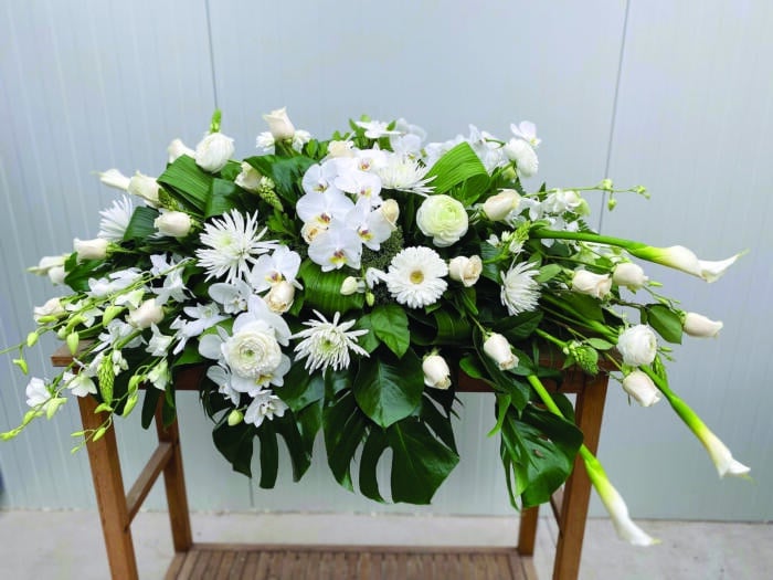 The Watering Can | An all white casket spray made with calla lilies, roses, dendrobium orchids, mums, star of Bethlehem, ranunculus, orchids, and trachelium in a bed of tropical greens.