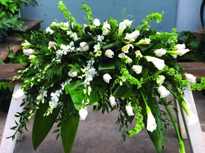 The Watering Can | An all white casket spray made with calla lilies, bells of Ireland, white roses, dendrobium orchids, freesia in a bed of lush greenery.