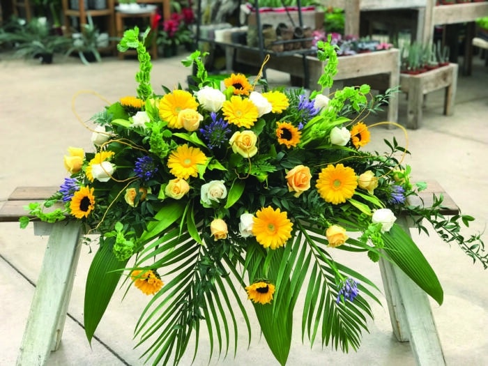 The Watering Can | A casket spray made with purple agapanthus, bells of Ireland, sunflowers, yellow gerberas, soft yellow roses, and white roses on a bed of greens.