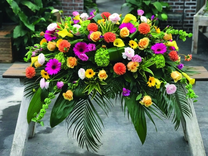 The Watering Can | A bright and colourful casket spray made with butterscotch roses, green hydrangea, yellow calla lilies, hot pink freesia, blush ranunculus, pink lisianthus, yellow gerberas, hot pink gerberas, orange ball dahlias, and bells of ireland on a bed of greens.
