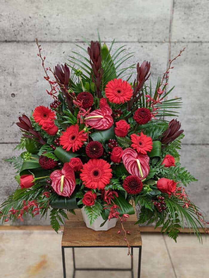The Watering Can | A deep red side spray made with roses, gerberas, leucadendron, dahlias, hypericum, annabelle orchids, and anthuriums backed by greens.