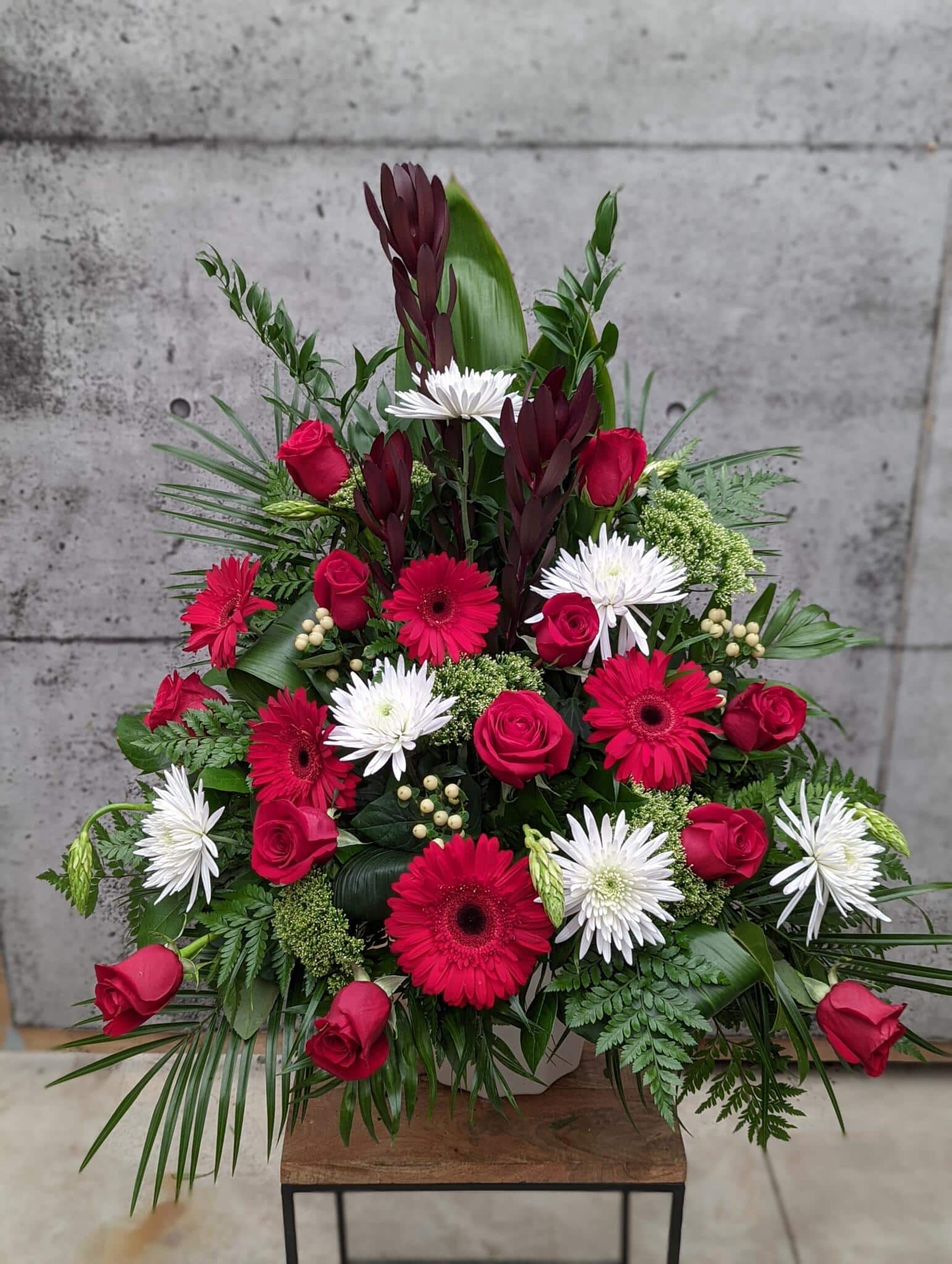 The Watering Can | A red and white side spray made with burgundy leucadendron, red roses, white mums, white trachelium, white hypericum, star of Bethlehem, and red gerberas set against a spray of greens.