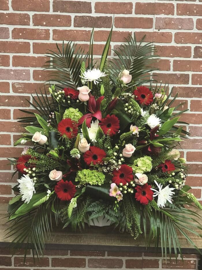 The Watering Can | A large side spray made with soft pink roses, white mums, burgundy lilies, burgundy gerberas, green hydrangea, stars of Bethlehem, white hypericum, soft pink freesia, white waxflower backed by greenery.