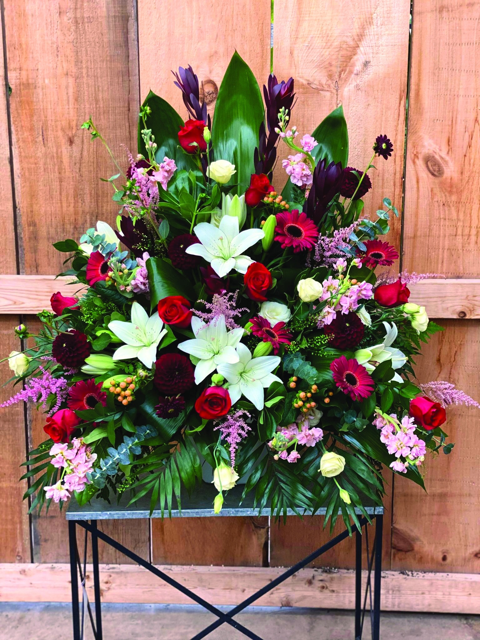 The Watering Can | A large side spray made with burgundy leucadendron, red roses, white lilies, blush stock, peach hypericum, soft pink astilbe, cream lisianthus, burgundy dahlias, and fancy greens.