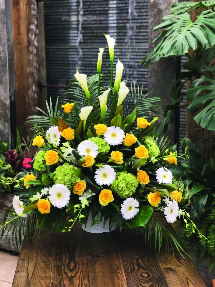the Watering Can | A yellow, white, and green side spray made with white calla lilies, green hydrangea, yellow roses, white gerberas, and white dendrobium orchids backed by greenery.
