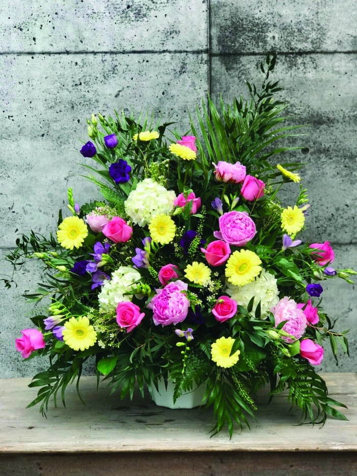 The Watering Can | A pink and yellow side spray made with white hydrangea, pink roses, lavender freesia, purple lisianthus, white waxflower, yellow gerbera daisies, and soft pink peonies backed by a variety of greens.