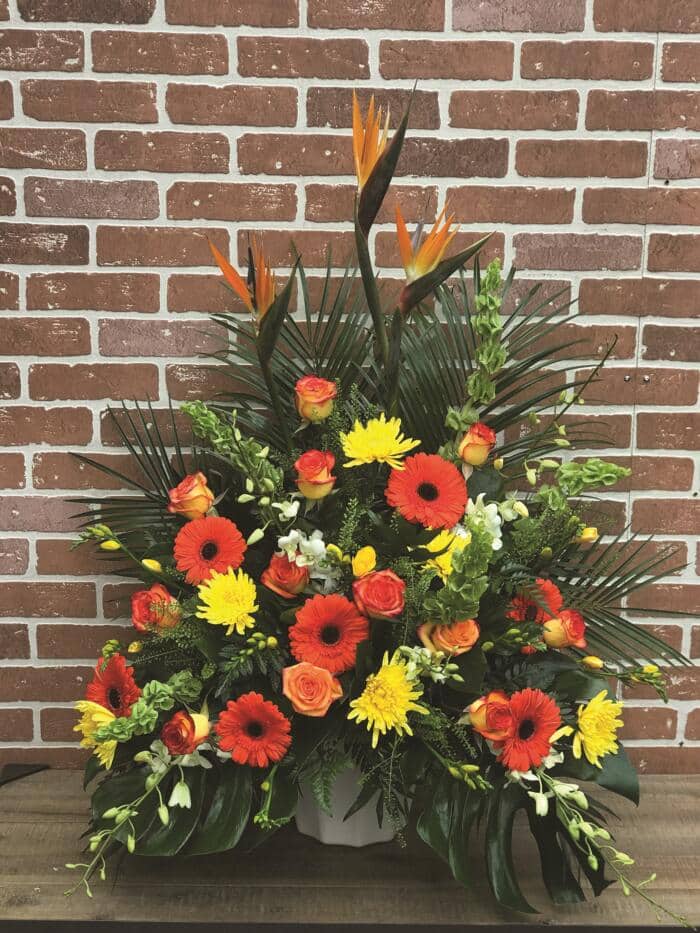 The Watering Can | A large orange and yellow side spray made with birds of paradise, bells of Ireland, yellow mums, orange roses, orange gerberas, yellow freesia, and white dendrobium orchids backed by greens.