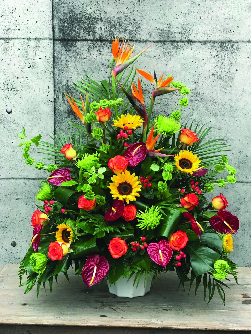 The Watering Can | A large bright side spray made with birds of paradise, bells of Ireland, sunflowers, green mums, burgundy anthuriums, orange roses, and red hypericum backed by greenery.