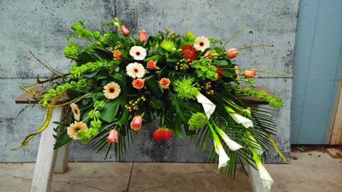 The Watering Can | A draping peach, and white casket spray made with white calla lilies, shimmer roses, bells of ireland, orange pincushion, peach gerberas, peach hypericum, white freesia, and green mums made in a lush bed of greens.