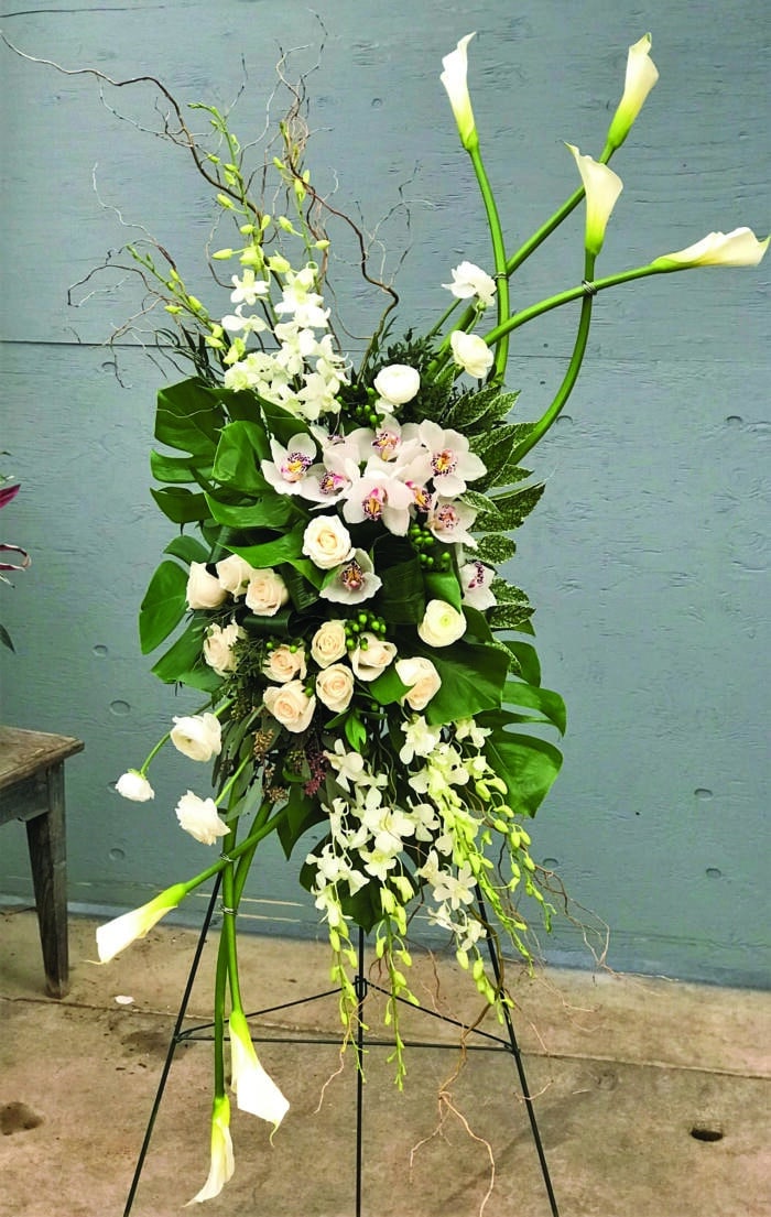 The Watering Can | An elegant white easel spray made with white calla lilies, white roses, stem white cymbidium orchid, white dendrobium orchids, green hypericum, white ranunculus, and tropical greens.