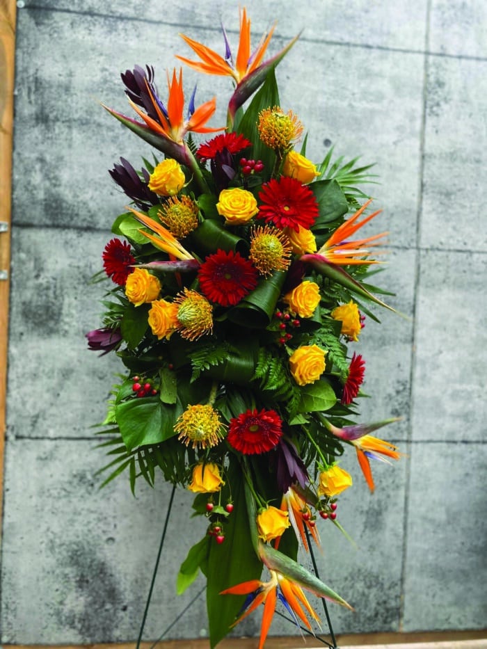 The Watering Can | A gorgoeus easel spray made with birds of paradise, yellow pincushion, yellow roses, red gerbera daisies, red hypericum, and burgundy leucadendron.
