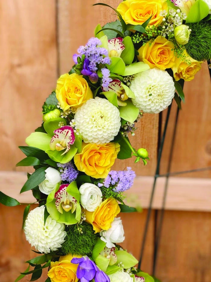 The Watering Can | Close up of the white dahlias, yellow roses, green cymbidium orchids, purple freesia, and blue statice on a floral funeral wreath.