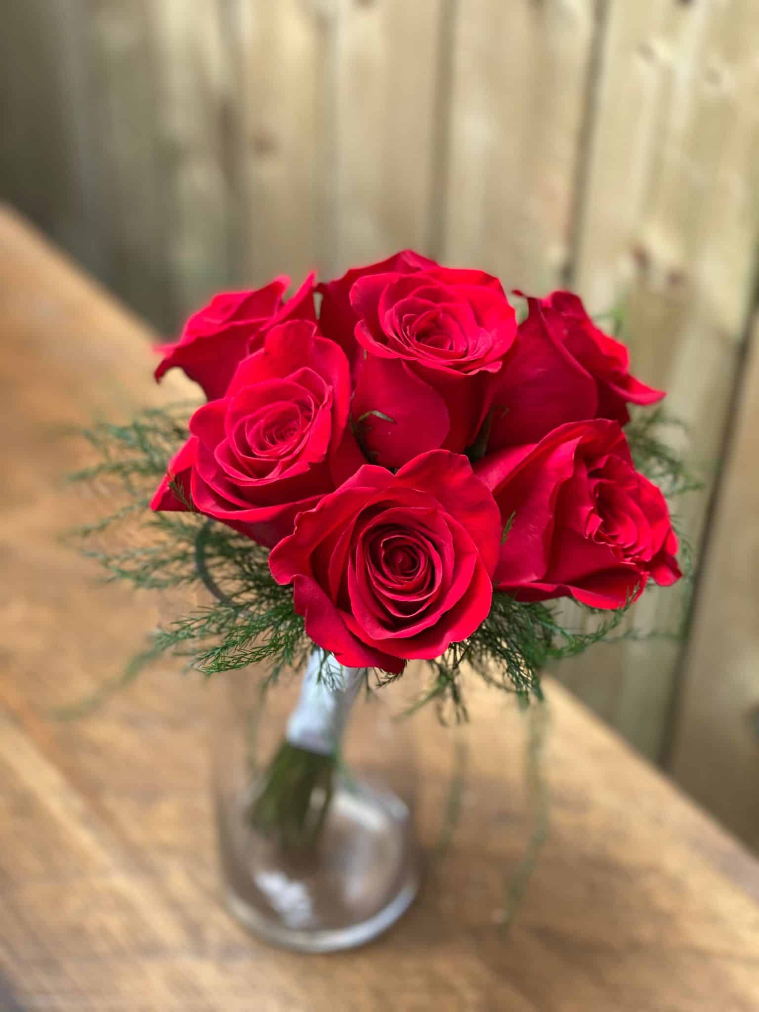 The Watering Can | a bouquet of 7 red roses with delicate tiki fern around the outside and white ribbon wrapped around the stems in a glass vase.