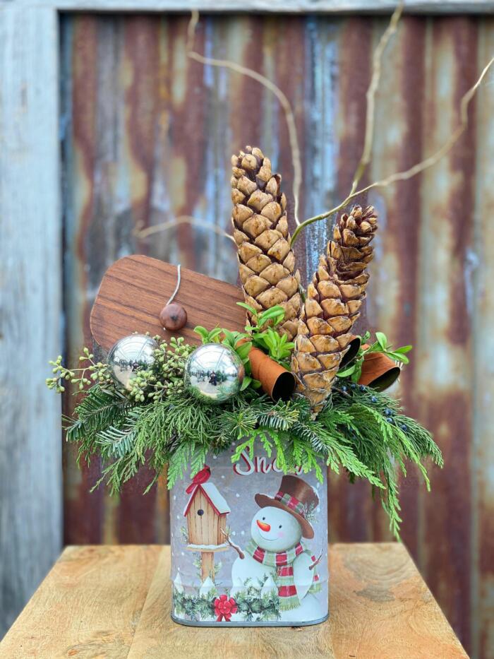 The Watering Can | A festive arrangement with evergreens, pine cones, and silver balls in a tin container depicting a snowman and the word 'snow'.
