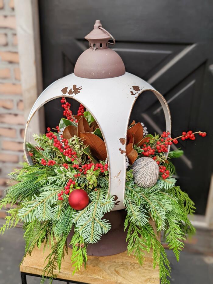 The Watering Can | A large round white lantern with a festive arrangment of evergreens, berries, and other red and white decorative elements spilling out on all four sides.