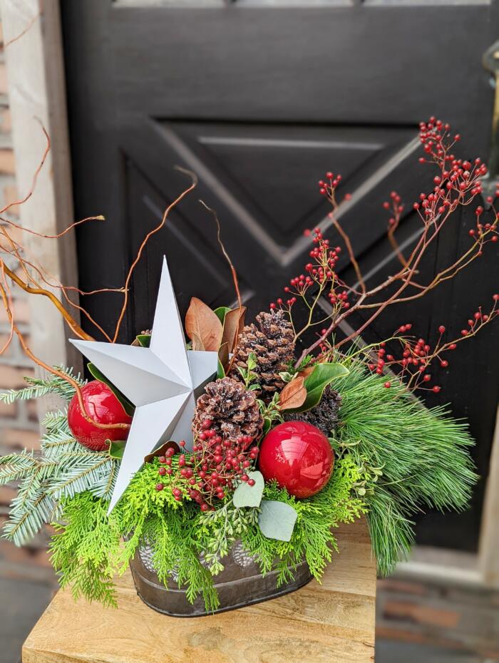 The Watering Can | A gorgeous festive arrangment of evergreens decorated with a large white star, natural pine cones, red balls, and rosehips in a oval tin container.