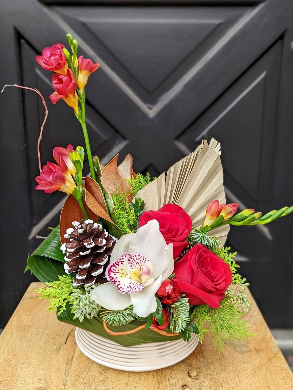The Watering Can | A European style arrangement with bright red roses and freesia, blush orchids, pine cones, and a palm spear amongst winter greens.
