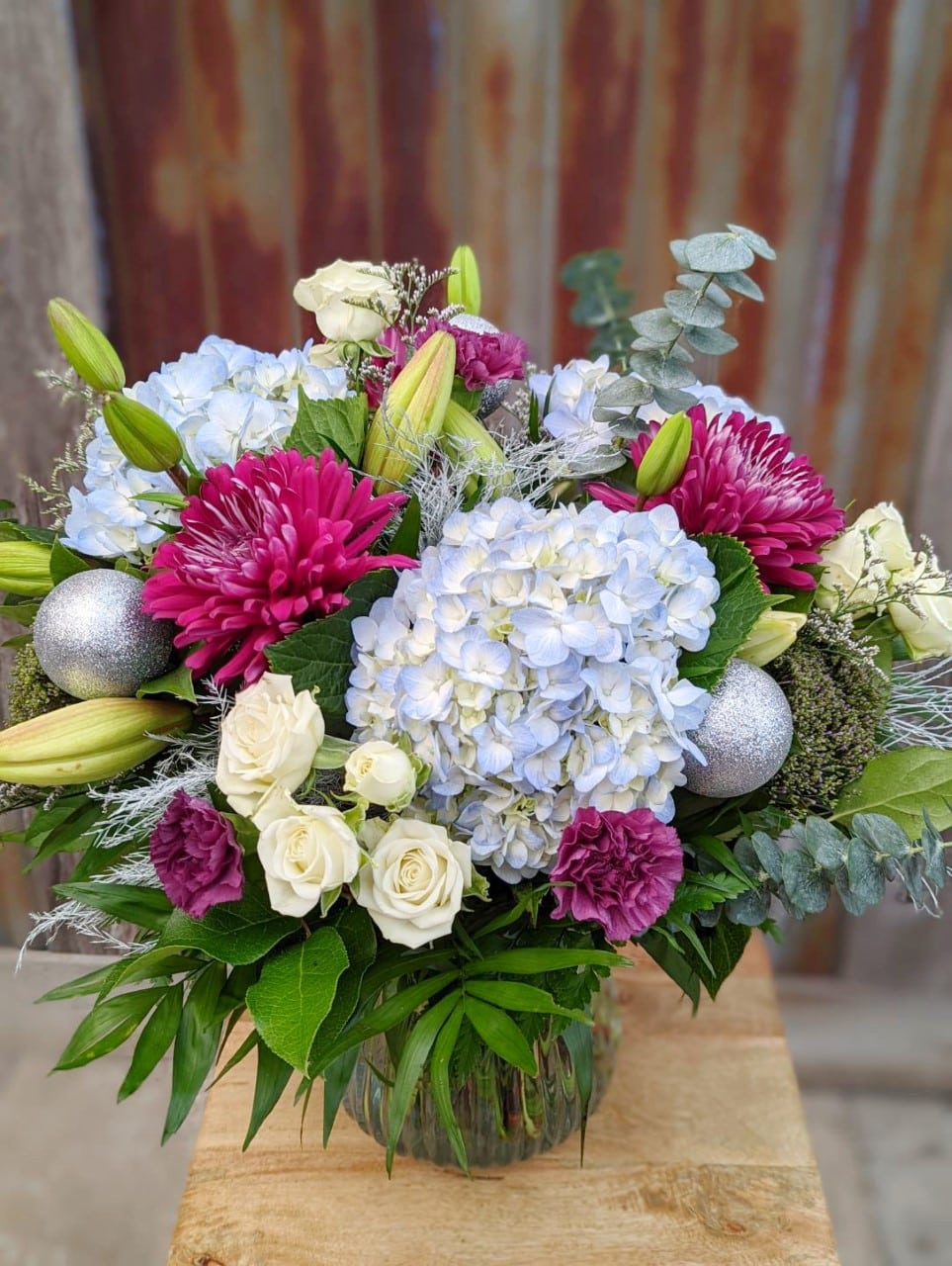 The Watering Can | A large bouquet made with plum, sky blue, and white blooms with silver balls in a blue glass vase.