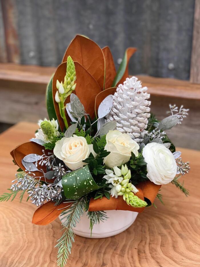 The Watering Can | A european style arrangement with white roses, star of bethlehem, magnolia, white ranunculus, and a white pine cone.
