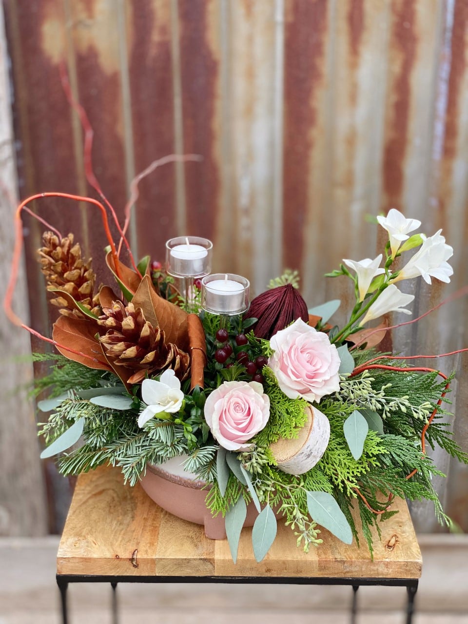 The Watering Can | A blush and burgundy European style arrangement with winter greens, pincones, and two tea light holders in a pink ceramic container.