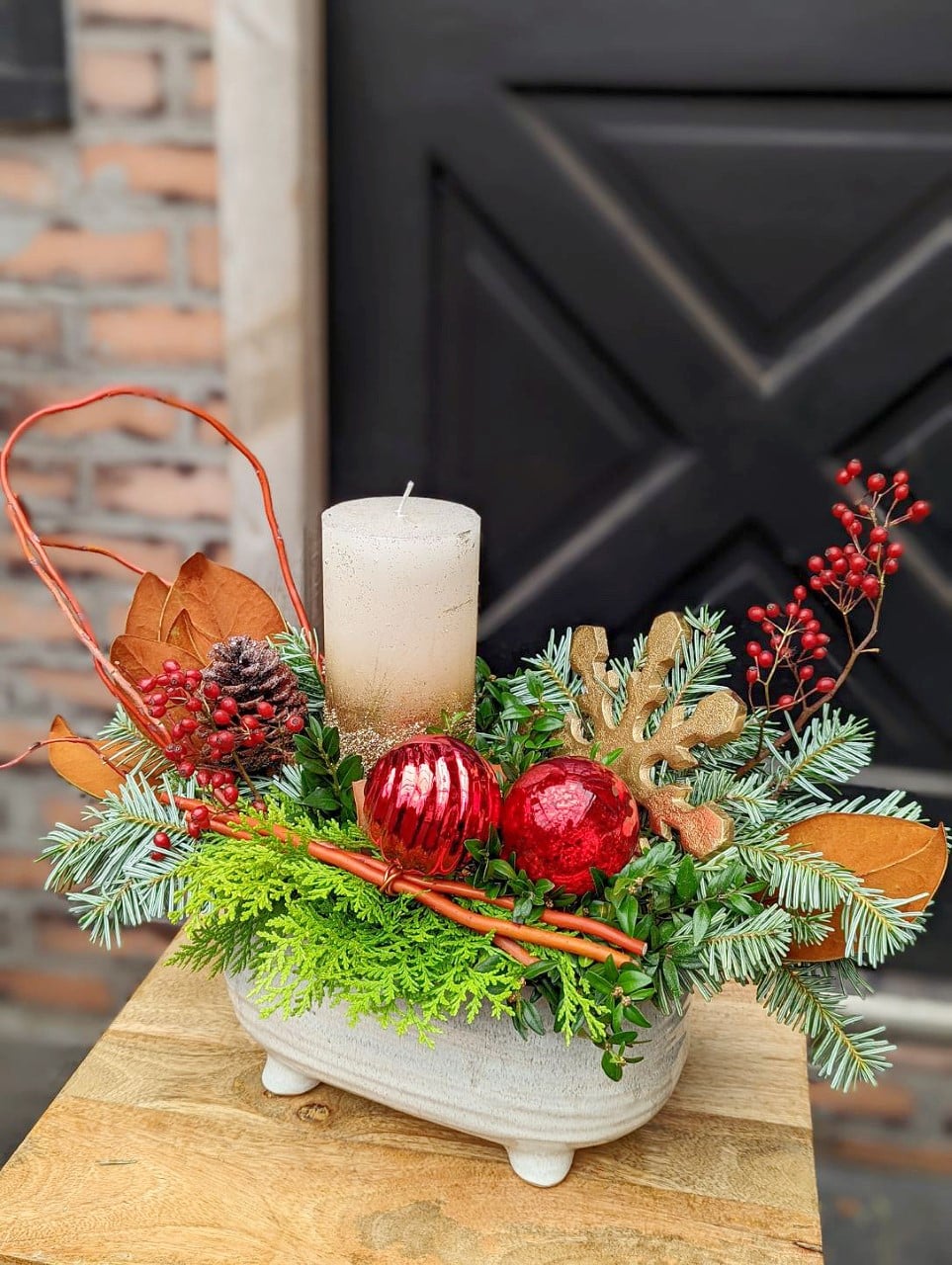 The Watering Can | A beautiful festive arrangement with a pillar candle to onse side, red natural deco with red accents in a white oval container.