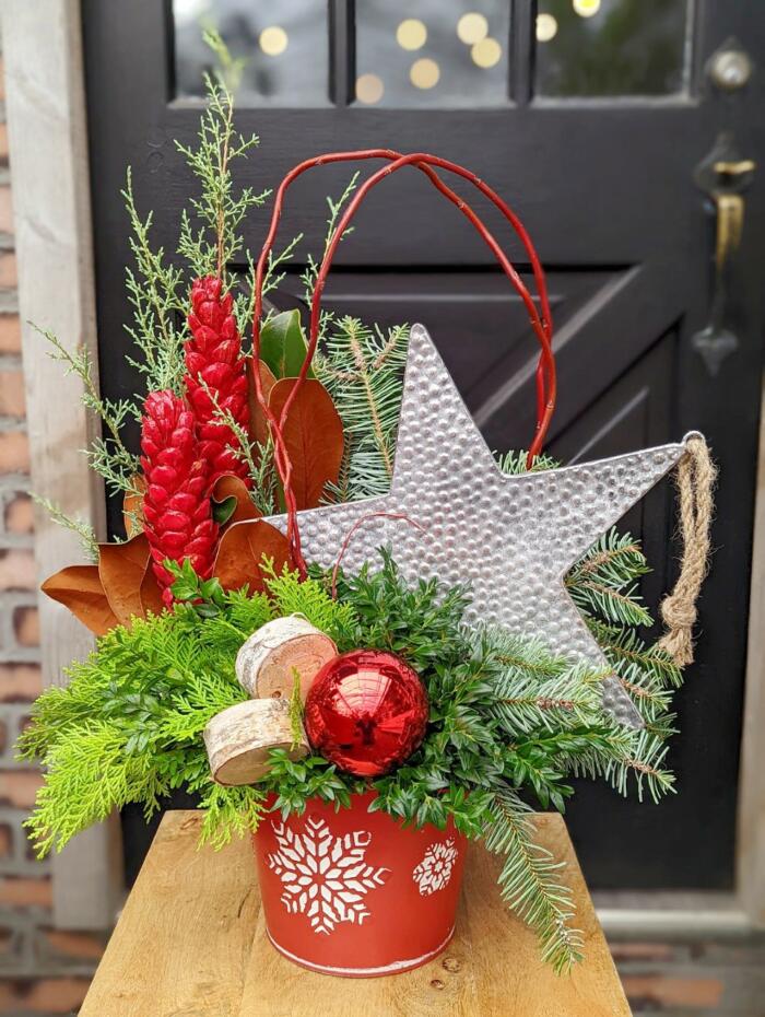 The Watering Can | A festive arrangement featuring a large silver star, two red strobus cones, a dogwood loop, red ball, and birch slices amongst winter greens in a round red container with a snowflake on it.